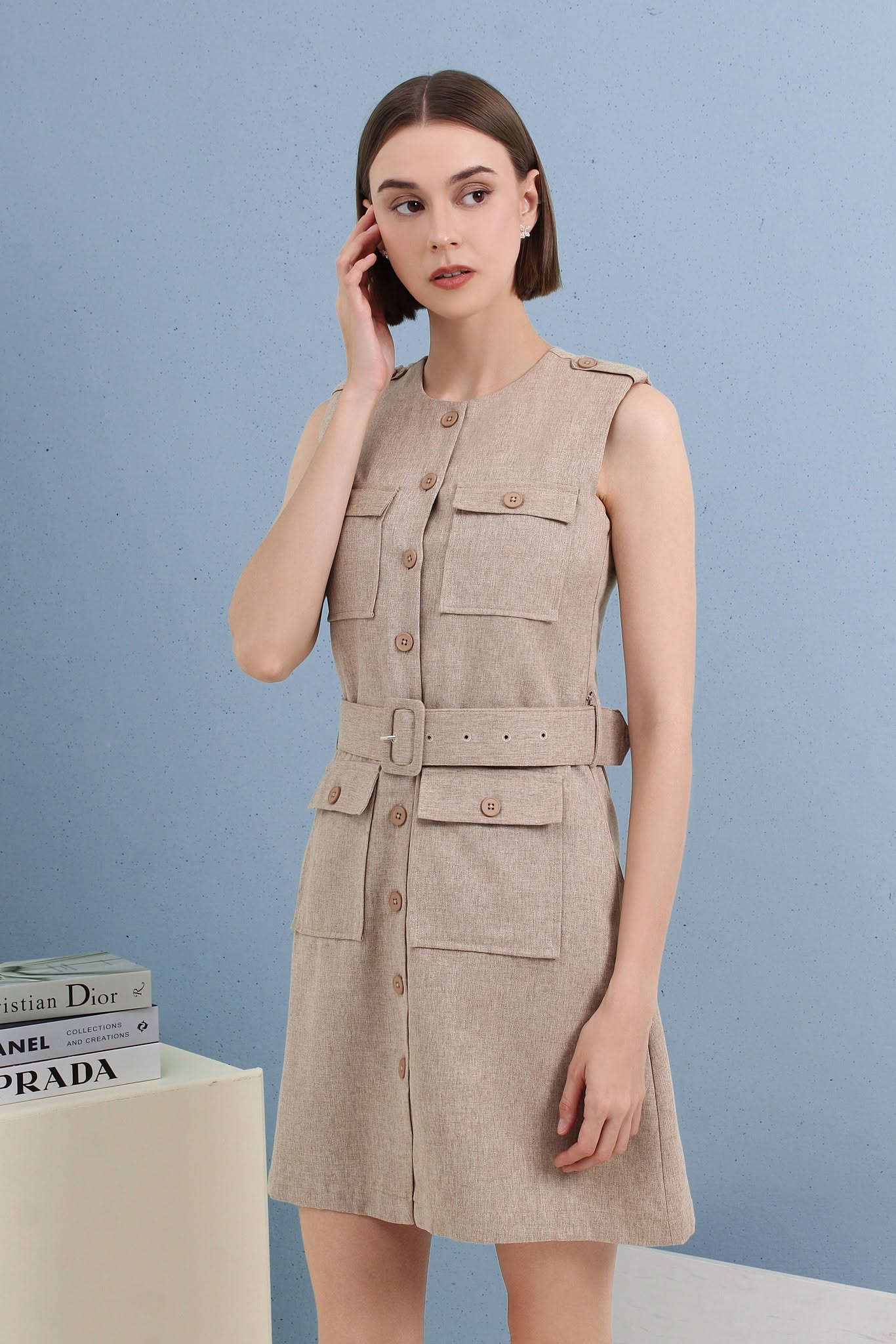 A woman modeling a sleeveless beige linen-look mini dress with a matching belt, button front, and flap pockets