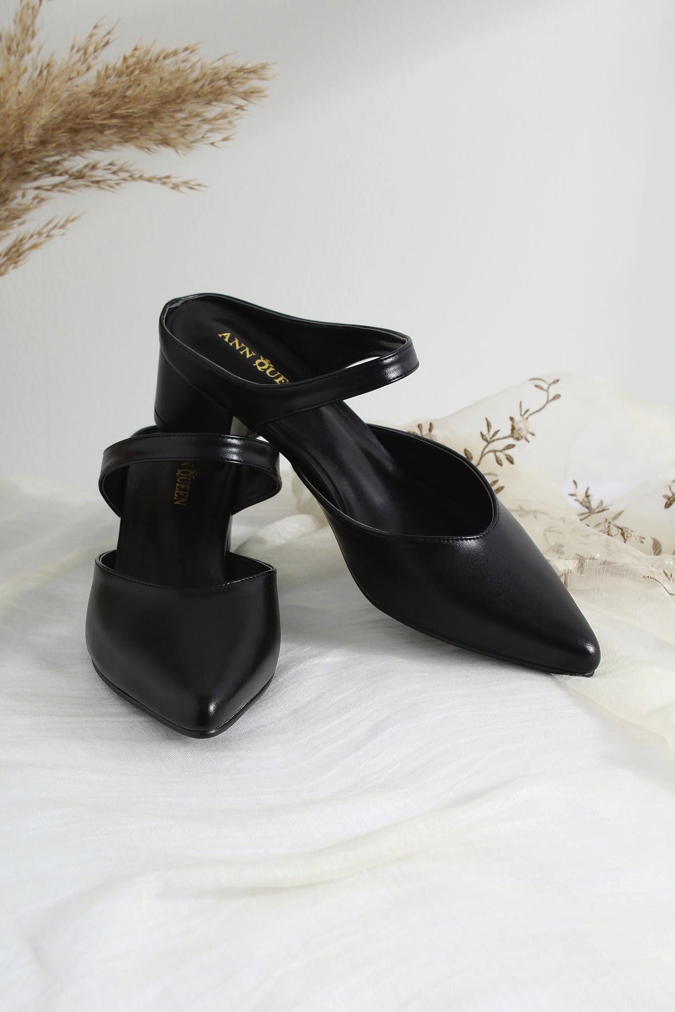 A pair of black pointed-toe mule heels with a low block heel and ankle strap, displayed on a white draped fabric surface with dried floral decor in the background.