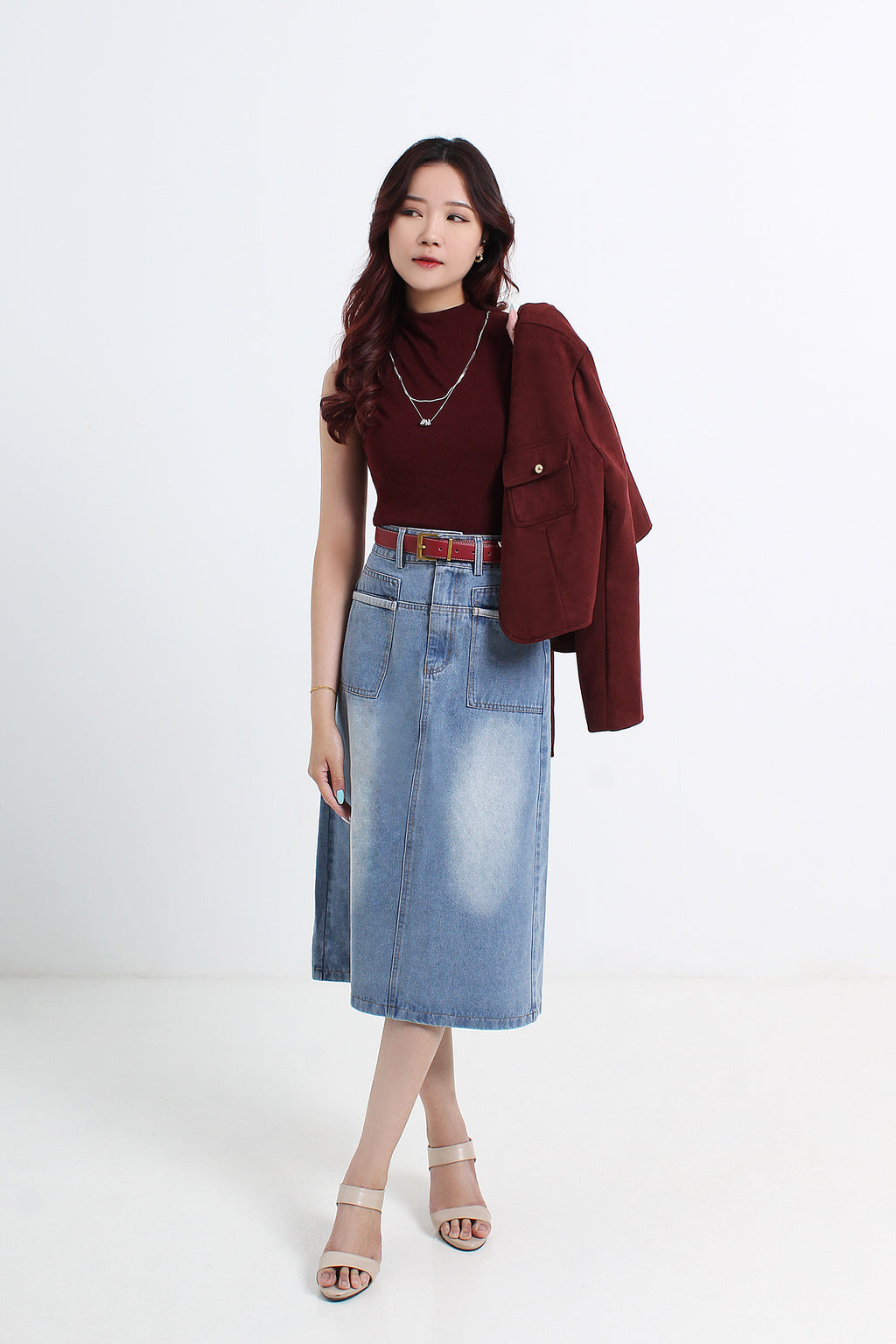 A woman standing and posing in a white top and light blue denim skirt with a belt, wearing dark-colored open-toe sandals.