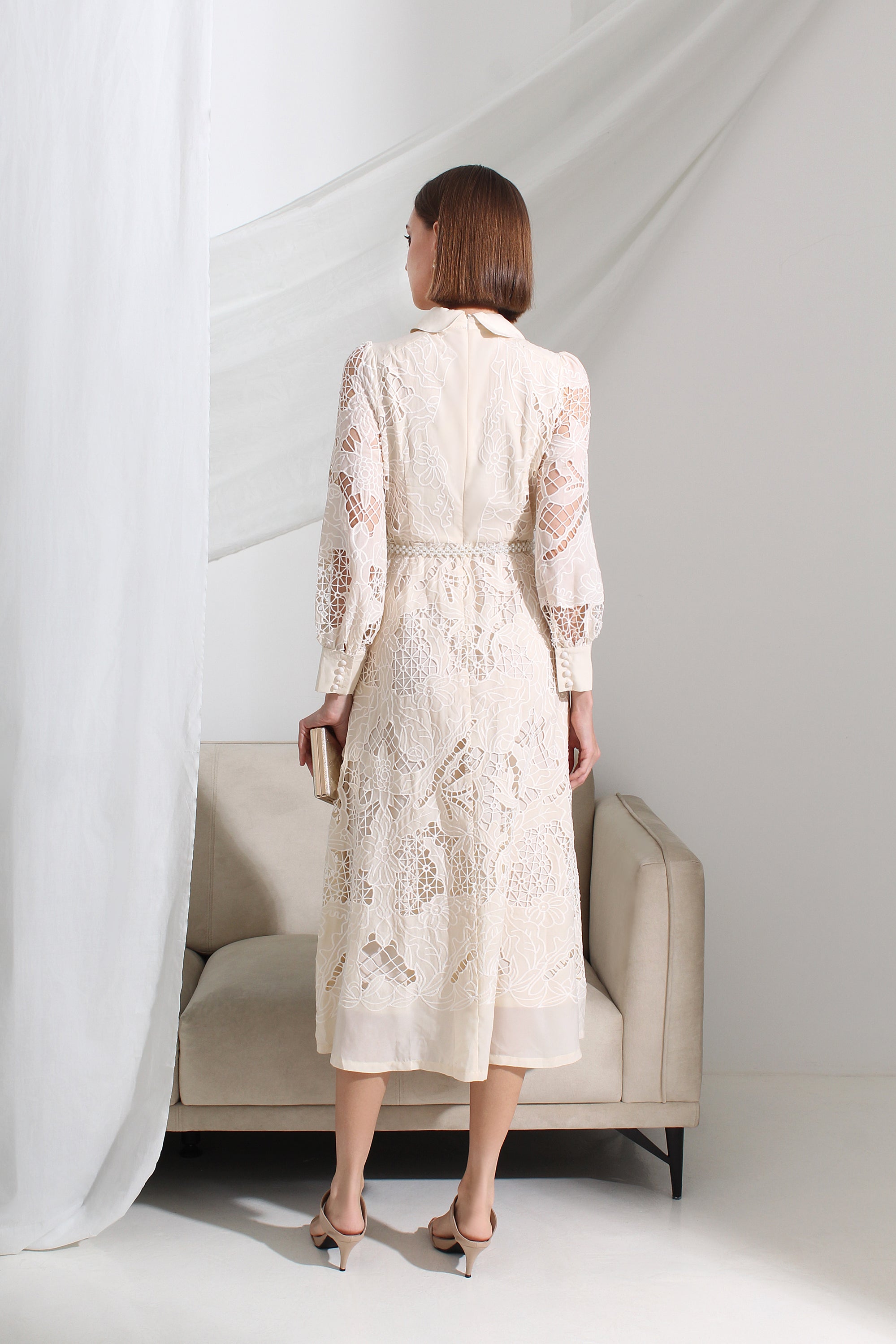 Woman in a white lace dress standing in a minimalistic room with a sofa and sheer curtains.