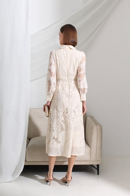Woman in a white lace dress standing in a minimalistic room with a sofa and sheer curtains.
