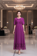 A woman wearing a purple floor-length evening gown with short puff sleeves, lace embroidery on the bodice, and a pleated skirt