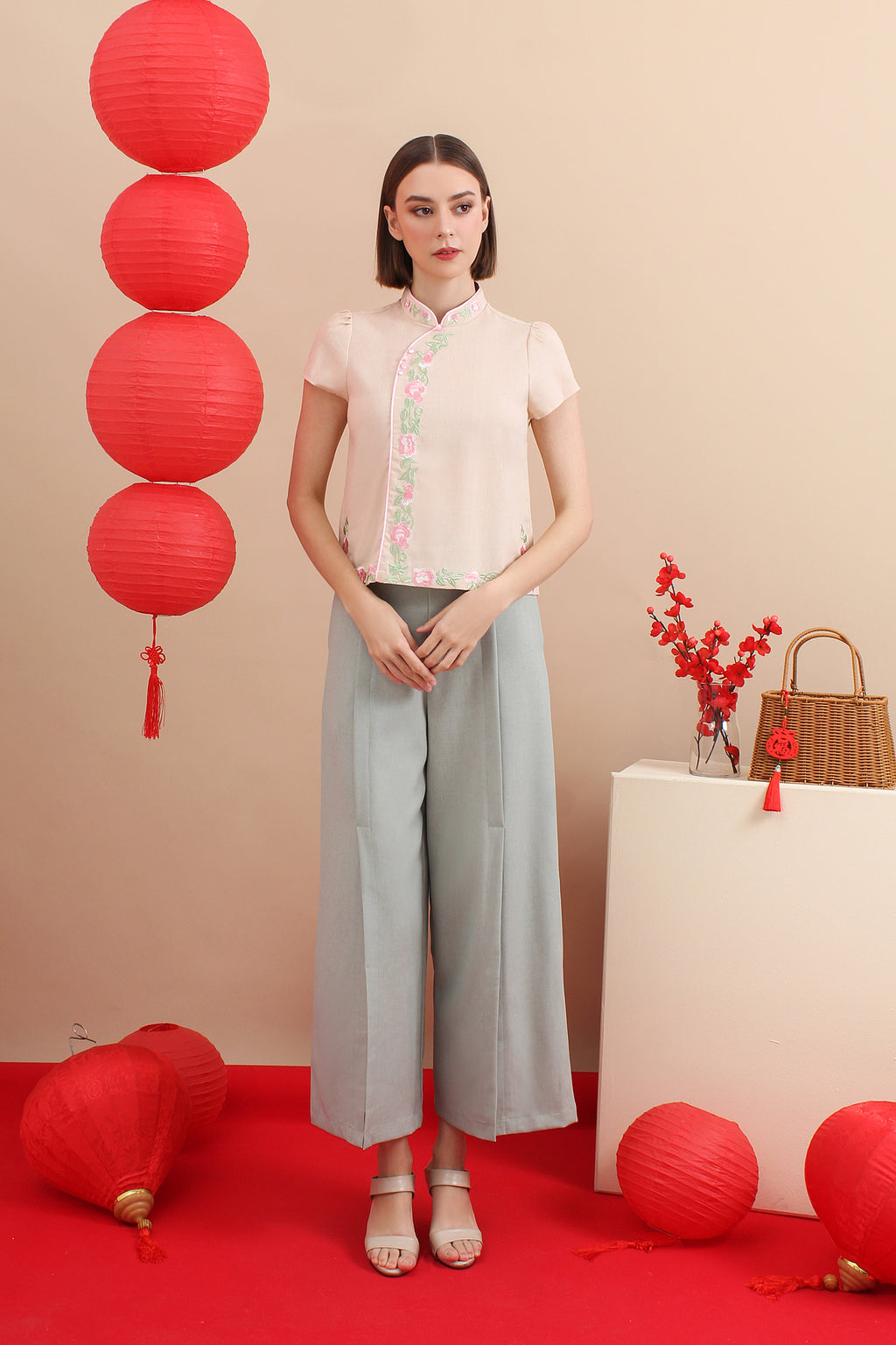 A female model wearing a pink and white cheongsam top with short sleeves and a mandarin collar, paired with gray trousers, standing in a staged setting with red decorative items and a handbag in the background