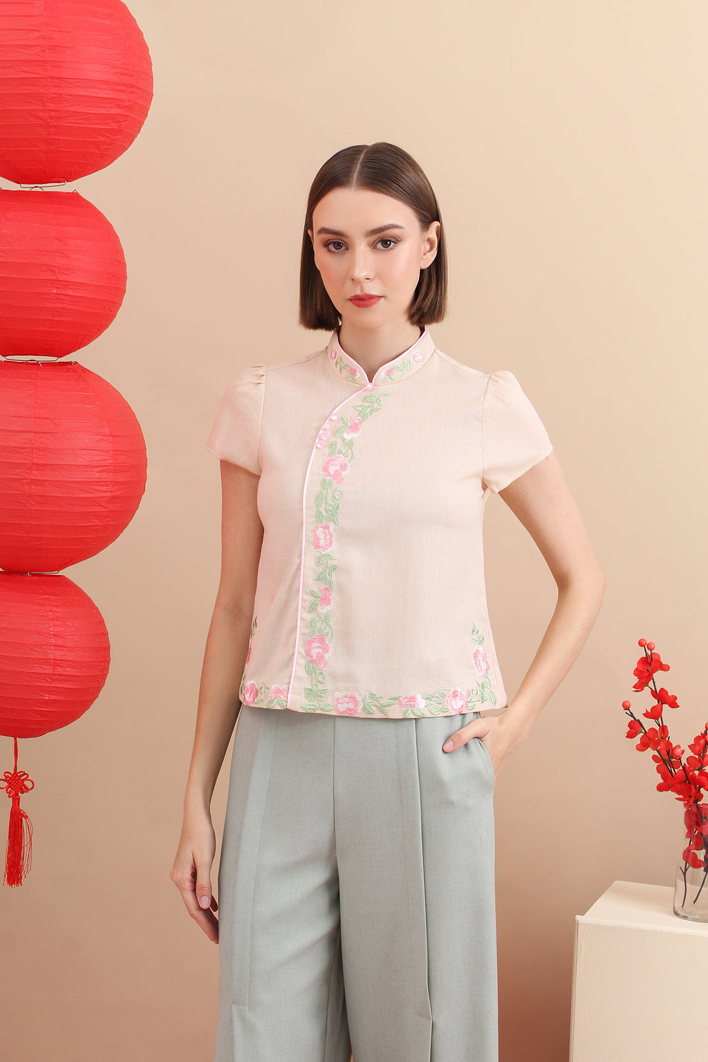 A woman standing in a room with red decorative balls and a floral-patterned pink cheongsam top with button details at the neckline, accompanied by matching gray trousers.