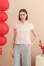 A woman standing in a room with red decorative balls and a floral-patterned pink cheongsam top with button details at the neckline, accompanied by matching gray trousers.