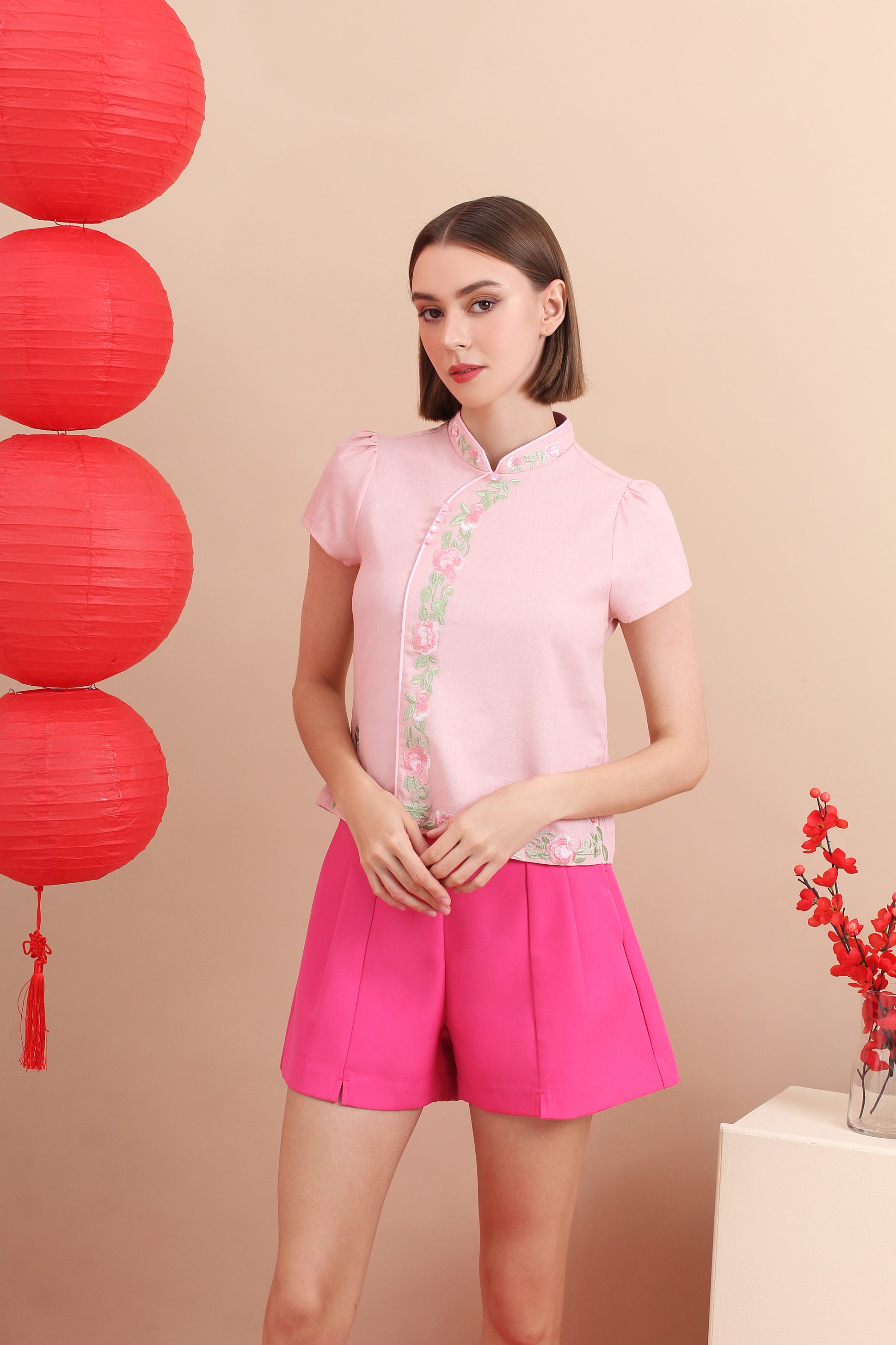 A woman standing in a room with two red lanterns hanging in the background, wearing a pink cheongsam mandarin collar top with floral patterns n it and pink shorts.