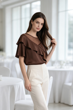 A woman modeling a brown ruffled short-sleeve blouse with a sheer V-neckline, paired with cream-colored trousers