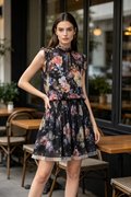 A woman models a sleeveless black floral dress with ruffled shoulders and a flared skirt