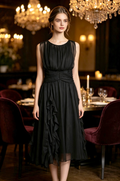 A woman stands in a dimly lit, elegant restaurant wearing a sleeveless black midi dress with a gathered bodice, sheer upper yoke, and layered ruffle skirt.