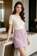 A woman stands indoors wearing a cream short-sleeve knit top and a light pink mini skirt with front button details and flap pockets.