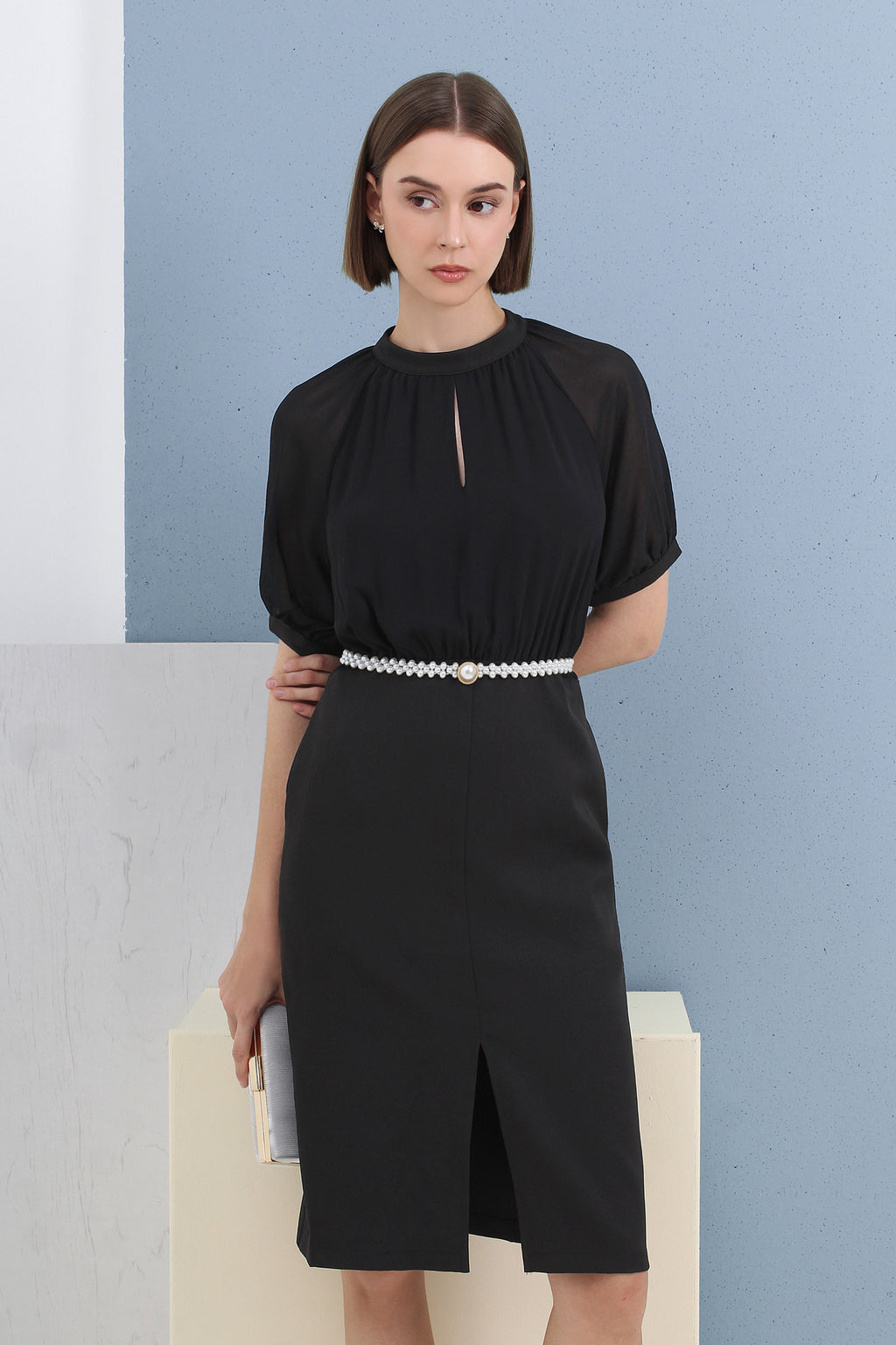 Woman wearing a black dress with a pearl belt against a light blue wall.