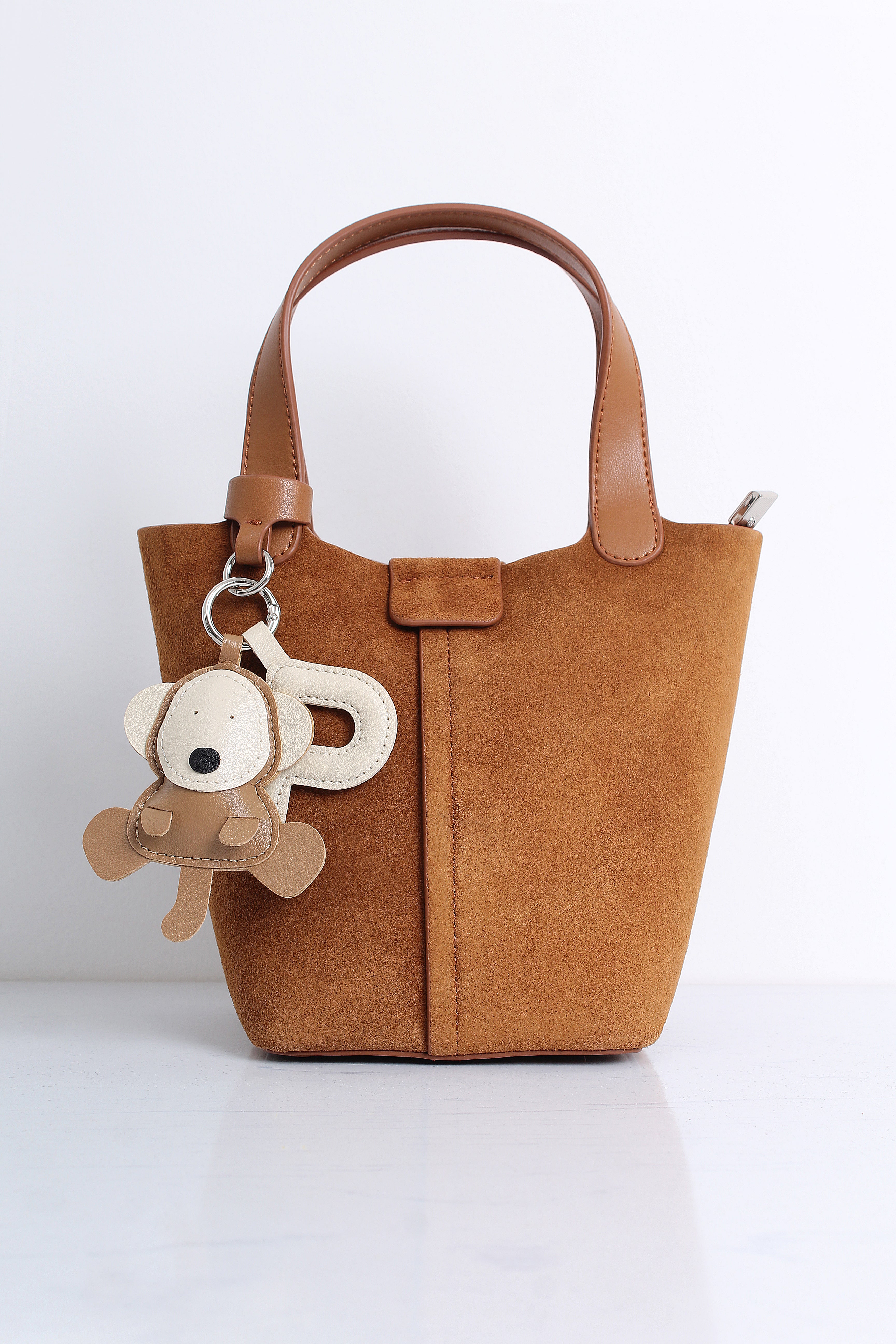 A small brown suede bucket bag with a top handle, featuring a hanging plush monkey keychain charm, set against a clean white background.