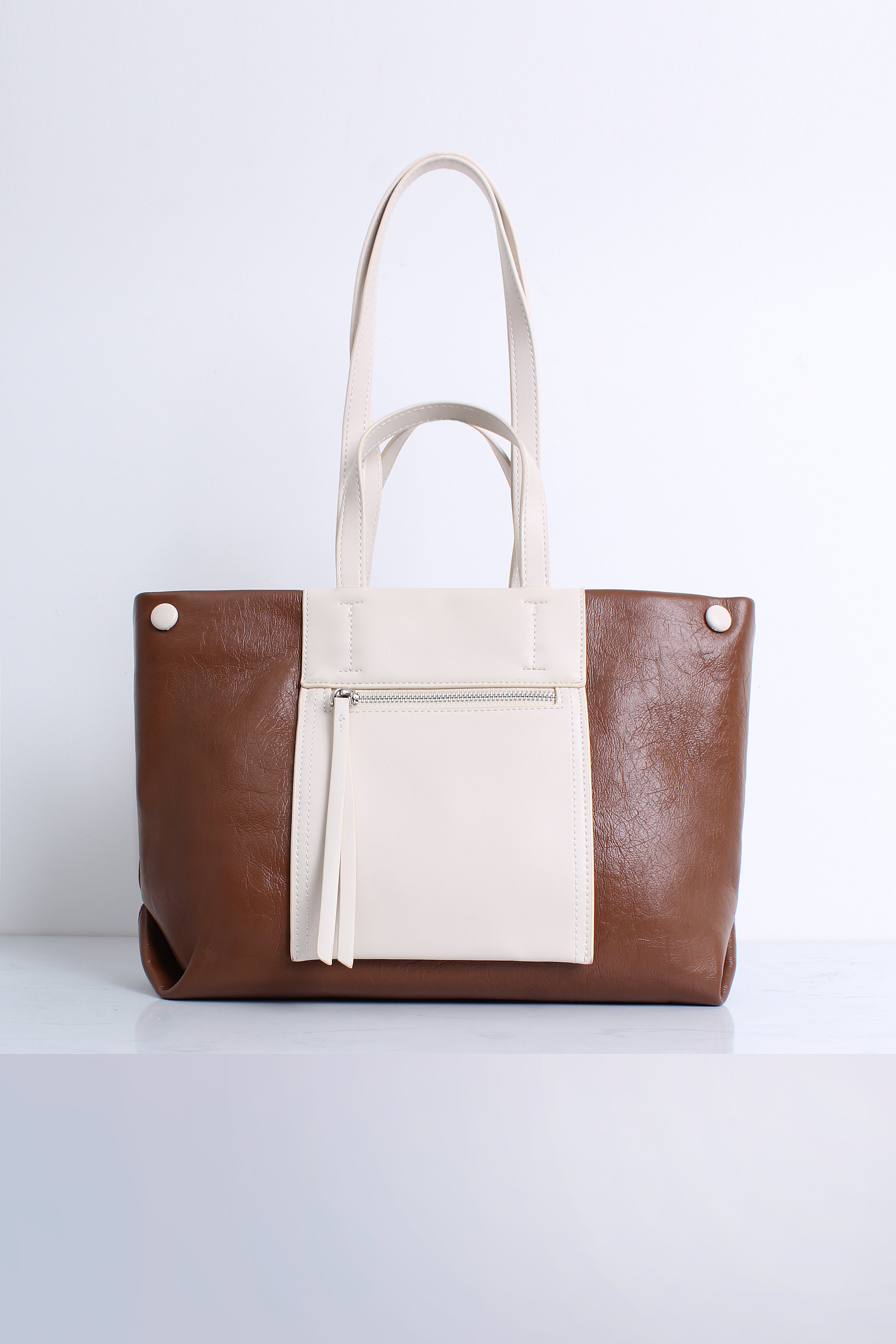 A structured tote bag in brown and cream faux leather, featuring dual cream shoulder straps, a front zip pocket with a tassel detail, and silver-tone hardware, set against a clean white background.