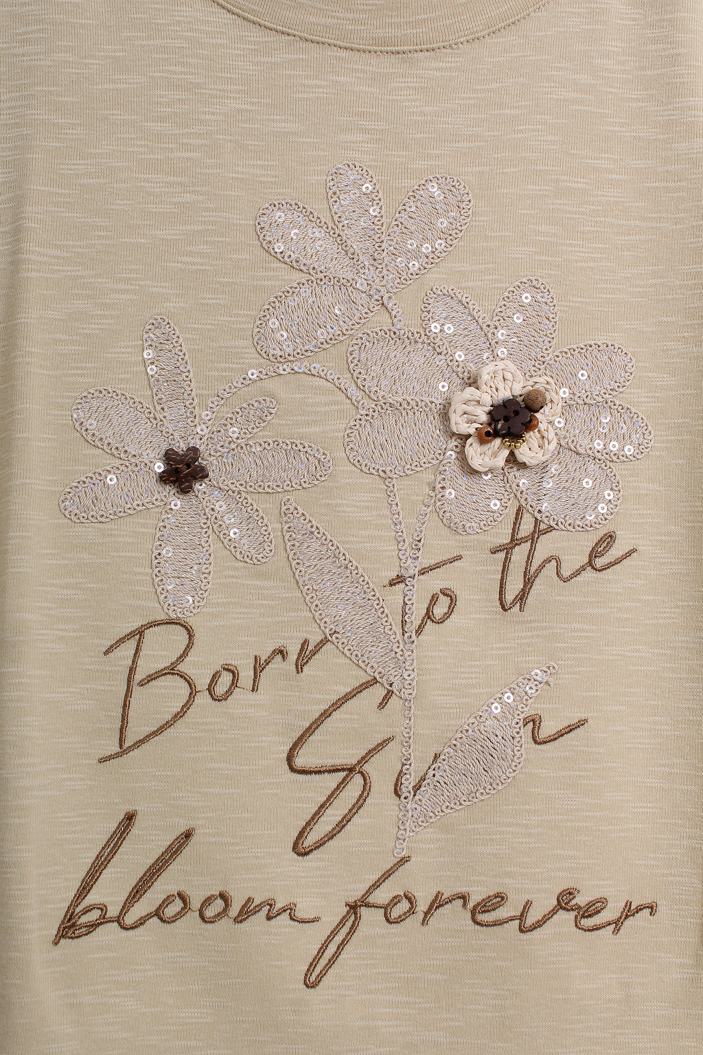 Close-up of a beige top featuring a sequined floral design and the embroidered text 'Born to the Sun bloom forever' in brown script.
