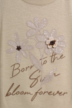 Close-up of a beige top featuring a sequined floral design and the embroidered text 'Born to the Sun bloom forever' in brown script.