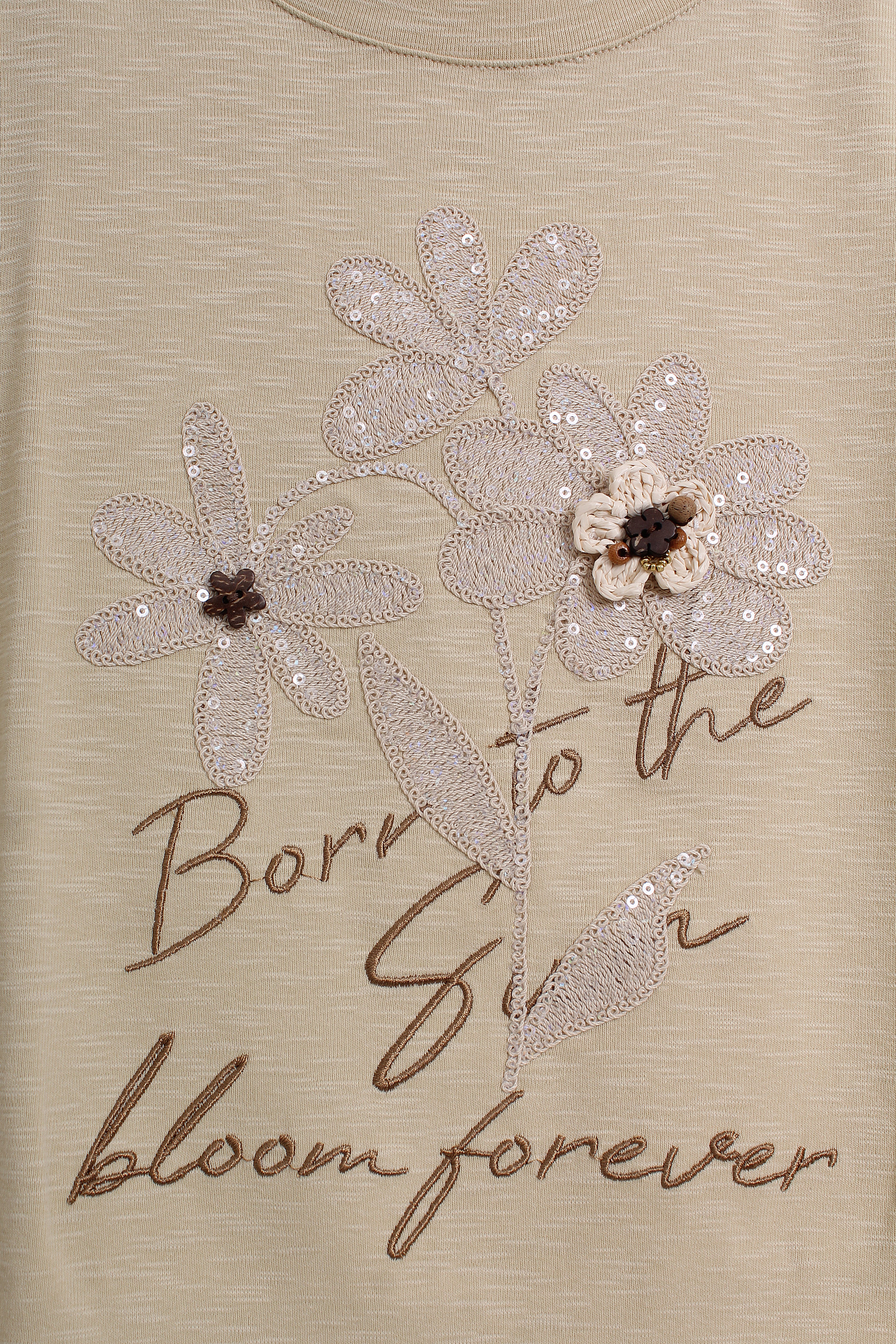 Close-up of a beige top featuring a sequined floral design and the embroidered text 'Born to the Sun bloom forever' in brown script.