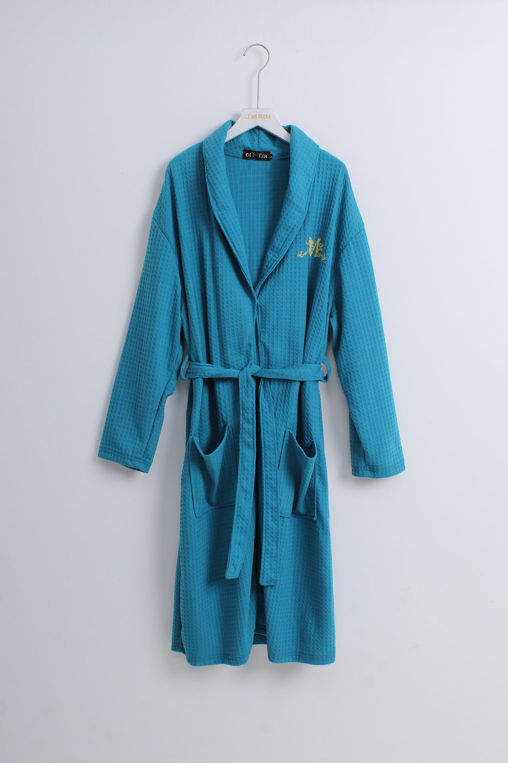A teal blue waffle-knit bathrobe hanging on a white hanger against a light gray wall. The robe features a shawl collar, a matching waist tie belt, front patch pockets, and a small gold embroidered monogram on the left chest.