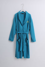 A teal blue waffle-knit bathrobe hanging on a white hanger against a light gray wall. The robe features a shawl collar, a matching waist tie belt, front patch pockets, and a small gold embroidered monogram on the left chest.