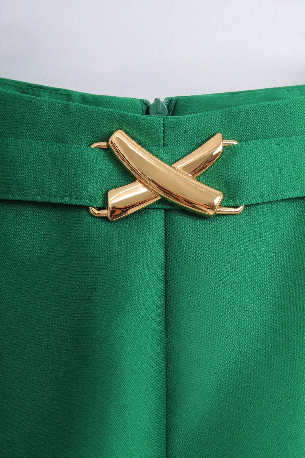 Close-up of a bright green skirt or trousers with a wide waistband and a prominent metallic gold cross-shaped belt buckle at the center.