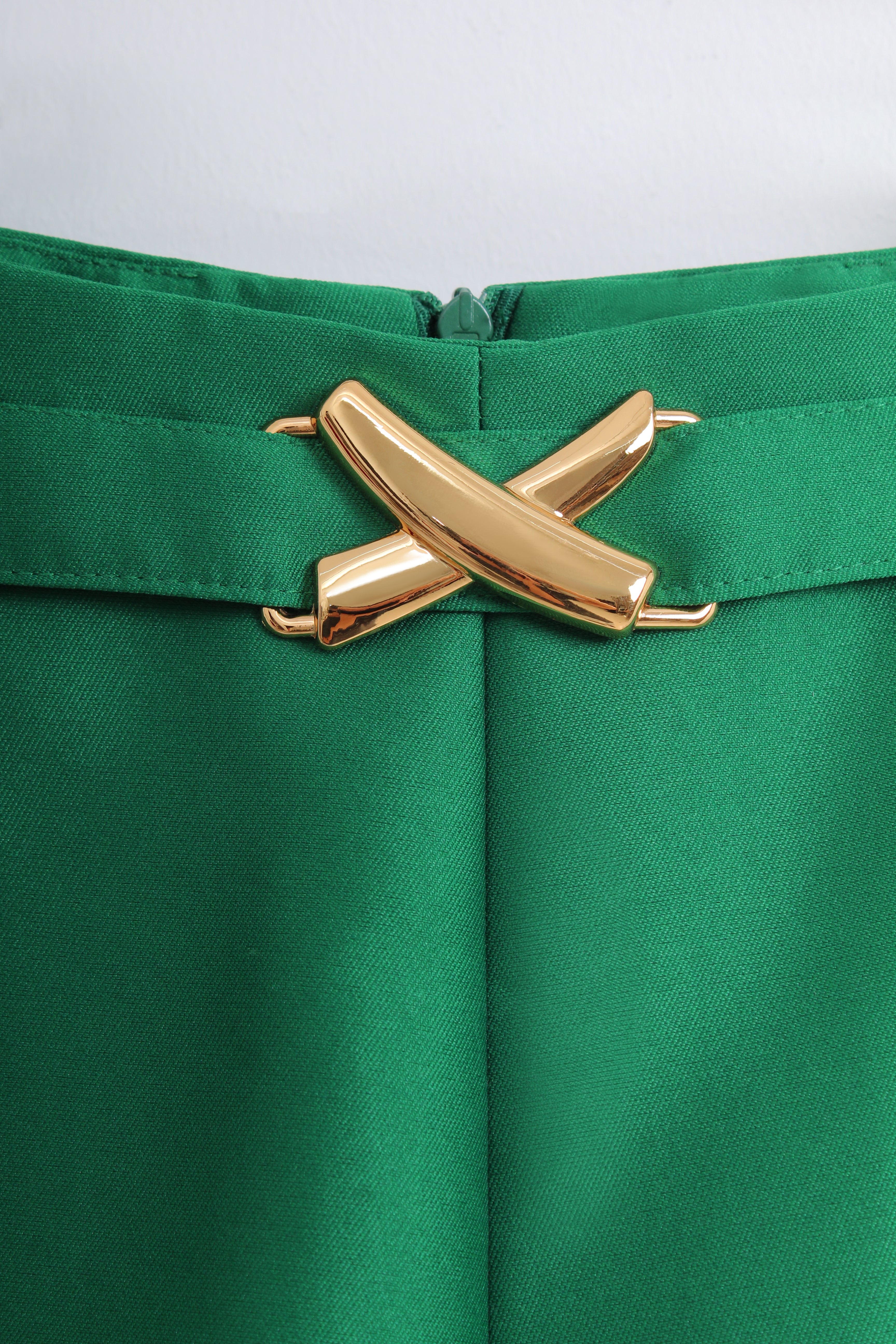 Close-up of a bright green skirt or trousers with a wide waistband and a prominent metallic gold cross-shaped belt buckle at the center.