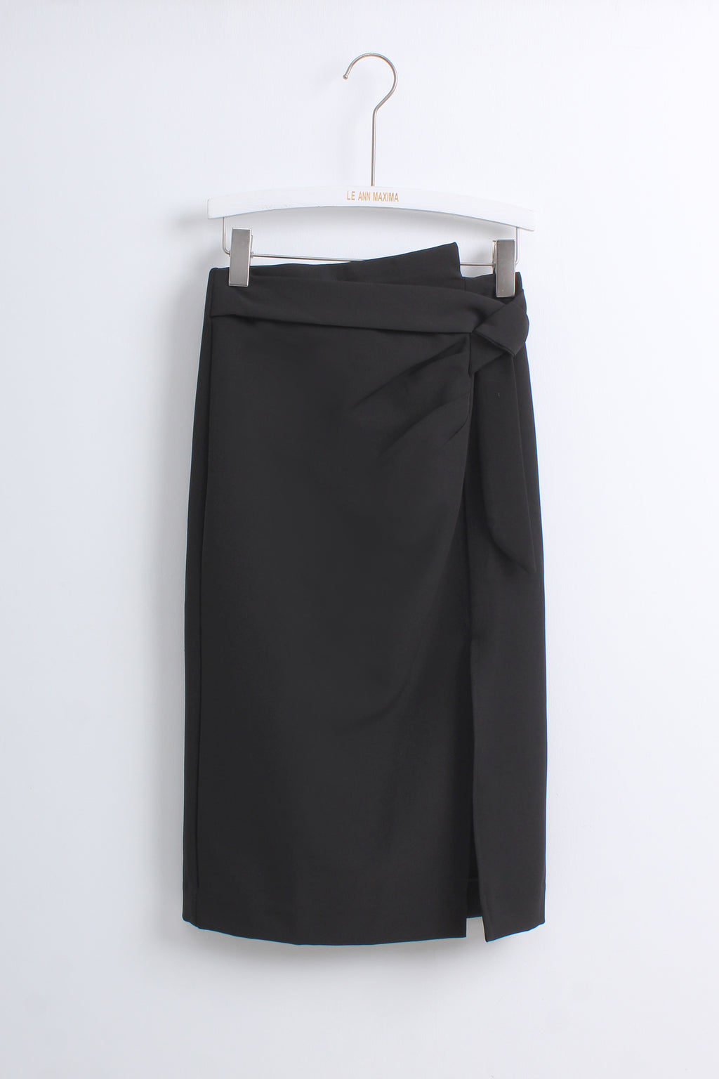 A black wrap-style pencil skirt with a side knot detail, hanging from a white hanger against a plain white wall.