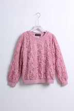 A pink textured lace knit crewneck sweater hanging on a white hanger against a plain light gray wall.