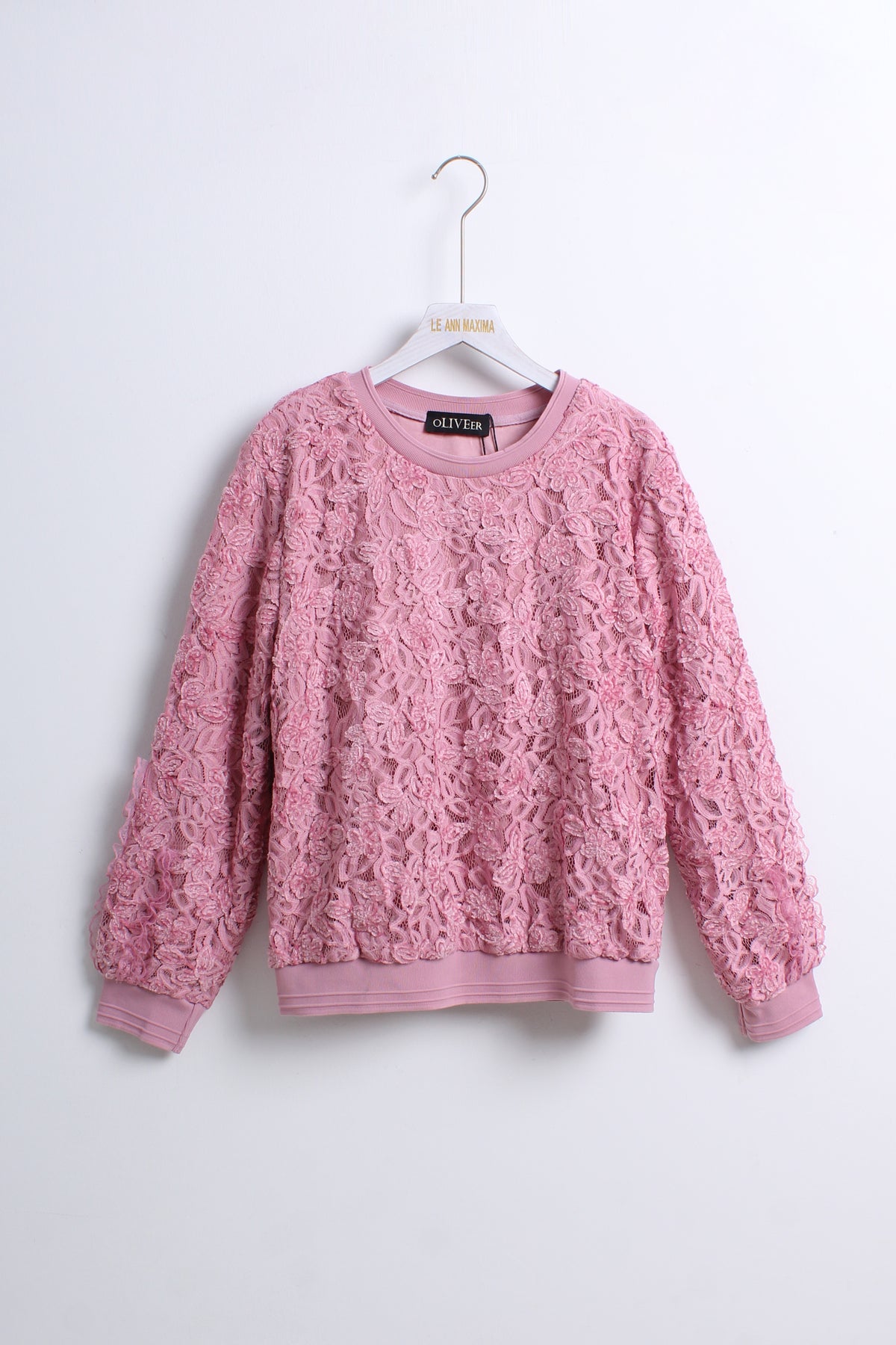 A pink textured lace knit crewneck sweater hanging on a white hanger against a plain light gray wall.