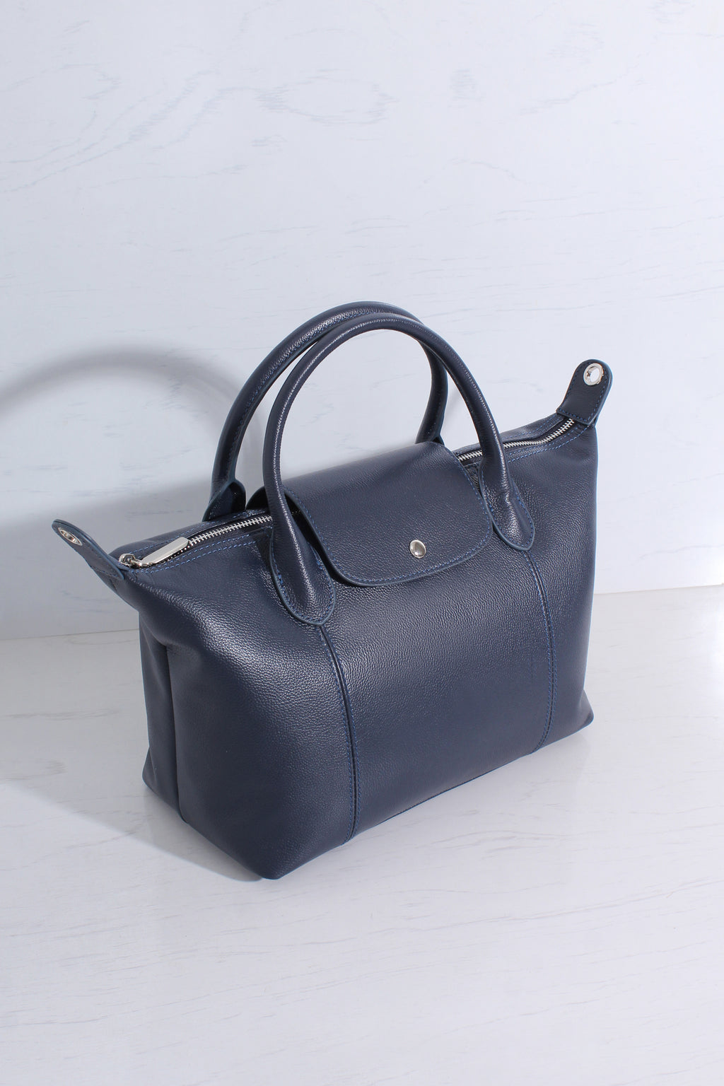 A dark navy blue leather handbag with double top handles and silver-tone hardware, photographed on a light gray surface against a matching light gray background.