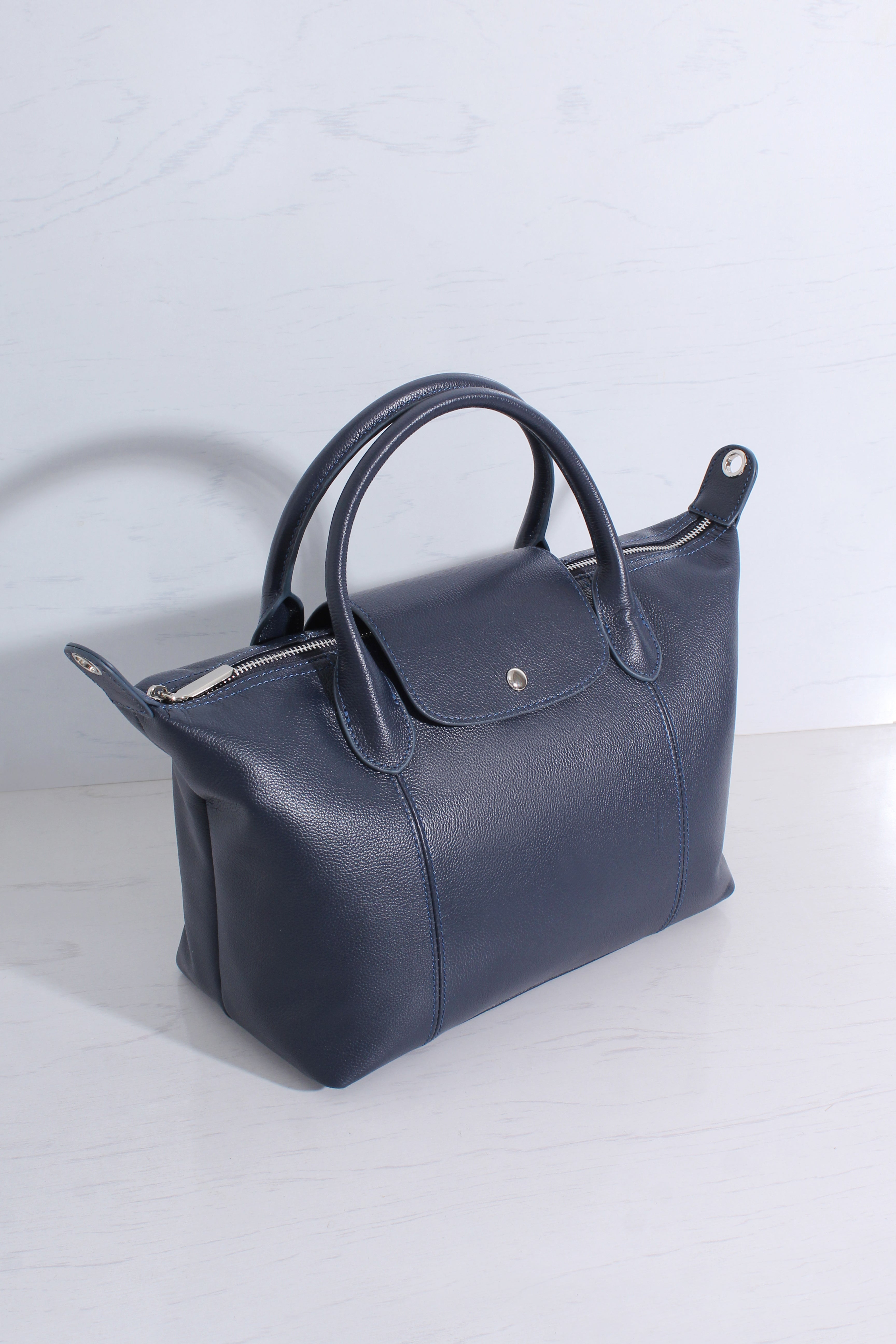 A dark navy blue leather handbag with double top handles and silver-tone hardware, photographed on a light gray surface against a matching light gray background.