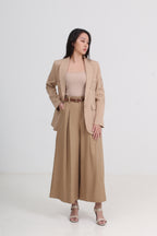 A woman models a beige two-piece suit consisting of a blazer with delicate lace embroidery on the lapels and sleeves, paired with matching wide-leg pleated trousers, a beige top, and brown leather sandals by Le Ann Maxima