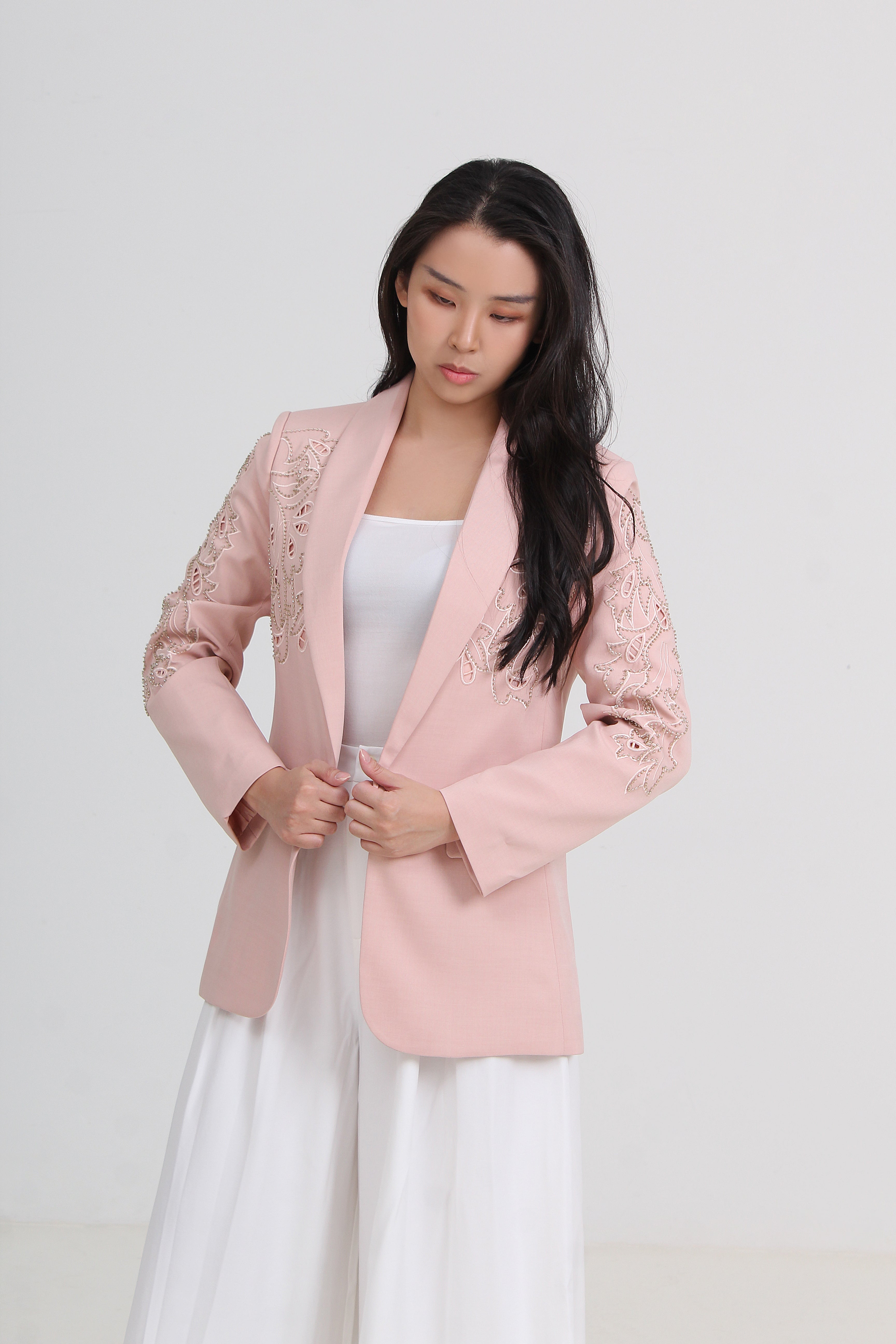 A woman wearing a light pink blazer with delicate floral embroidery on the sleeves, styled over a white top and white skirt