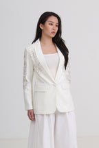 A woman modeling a white tailored blazer with delicate silver embroidery on the lapels and sleeves, paired with a white top and white wide-leg trousers by Le Ann Maxima
