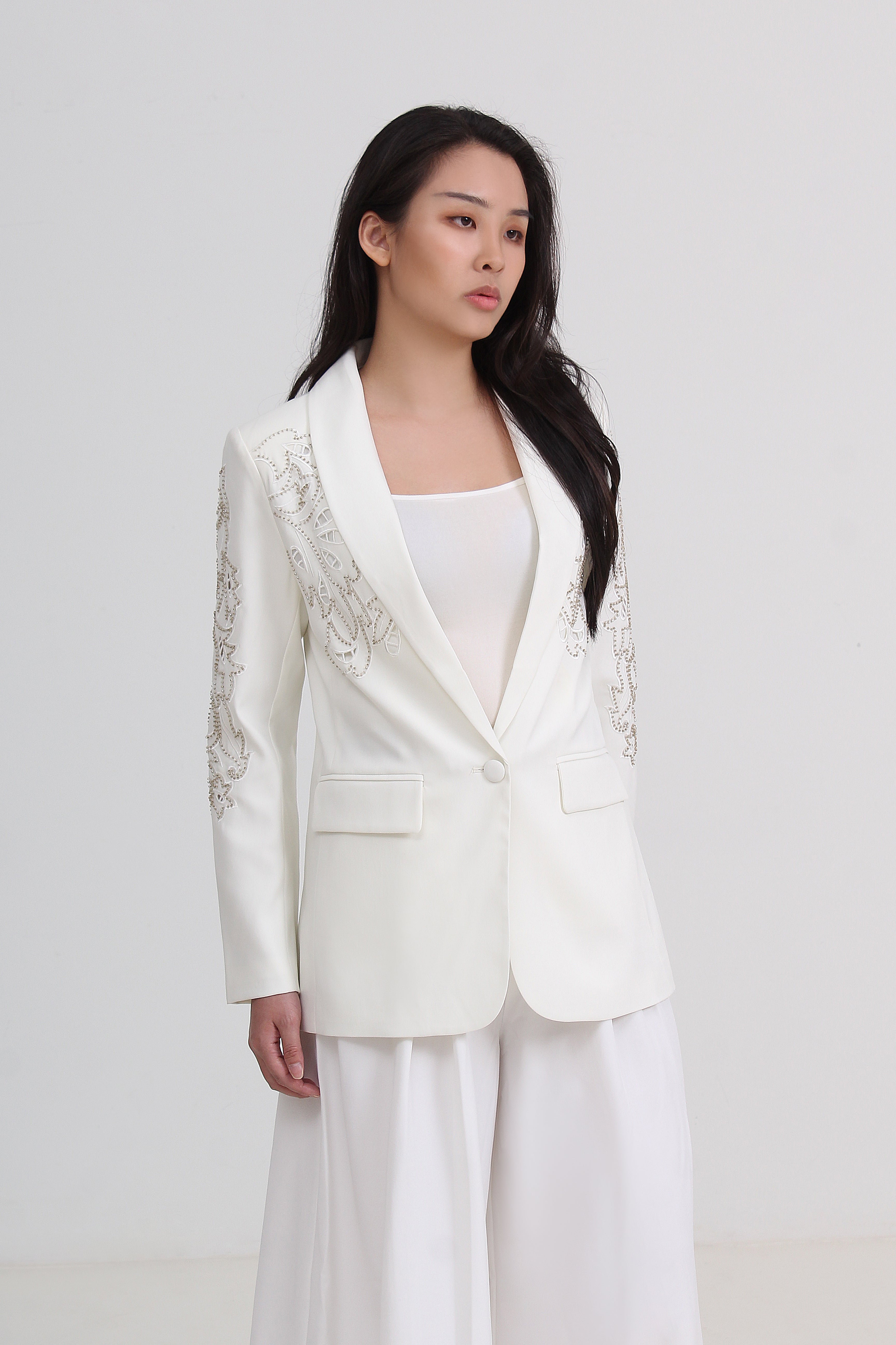 A woman modeling a white tailored blazer with delicate silver embroidery on the lapels and sleeves, paired with a white top and white wide-leg trousers by Le Ann Maxima