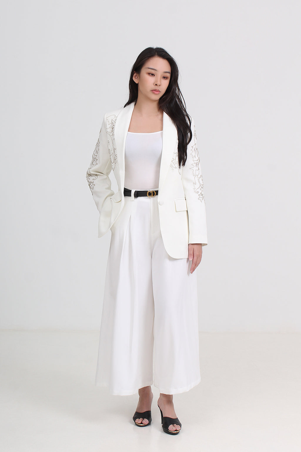 A woman modeling a white two-piece blazer and wide-leg pants set with a thin black belt, styled with a white top and black sandals by Le Ann Maxima