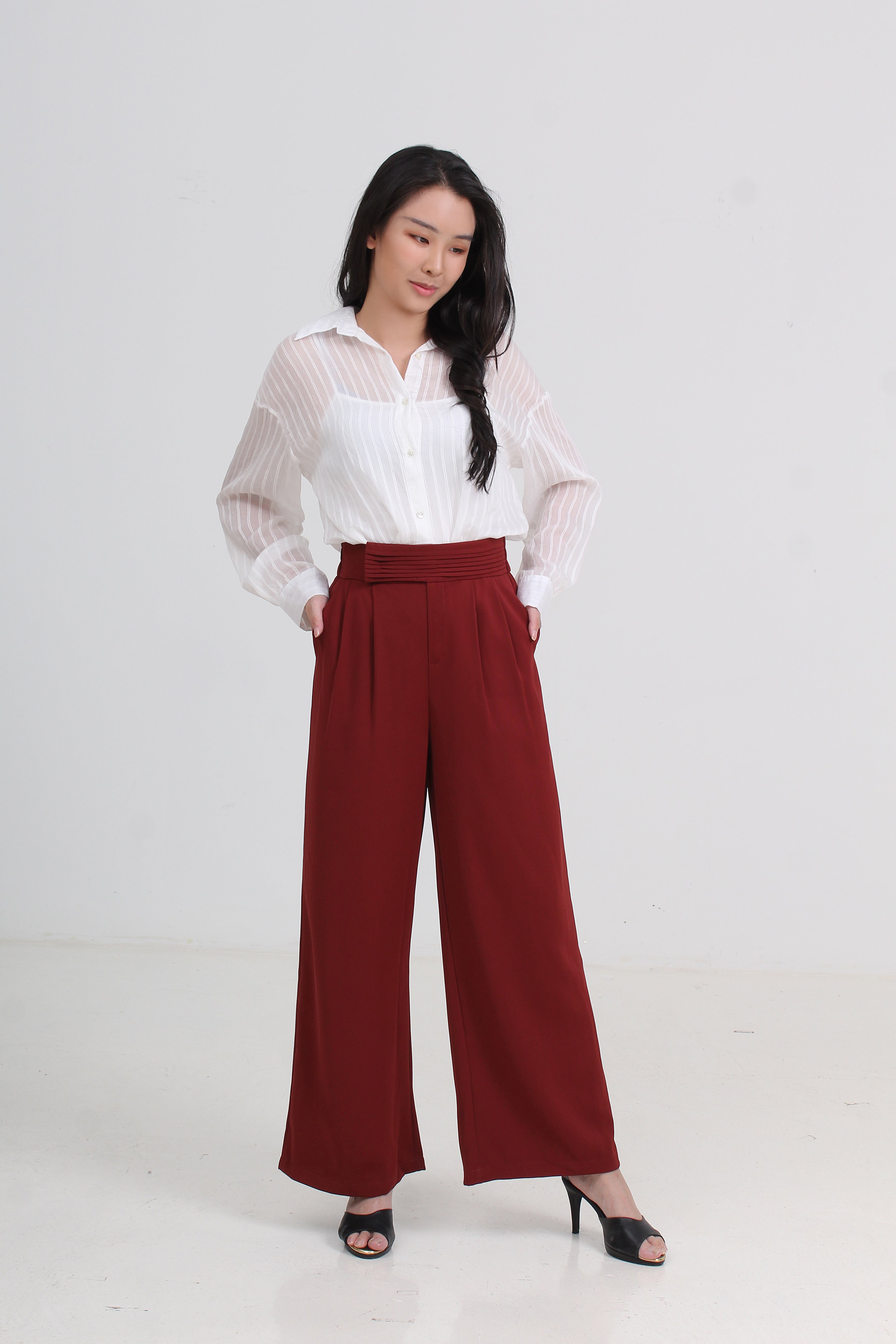 A woman modeling a white sheer long-sleeve button-up shirt paired with high-waisted, wide-leg burgundy trousers by Le Ann Maxima