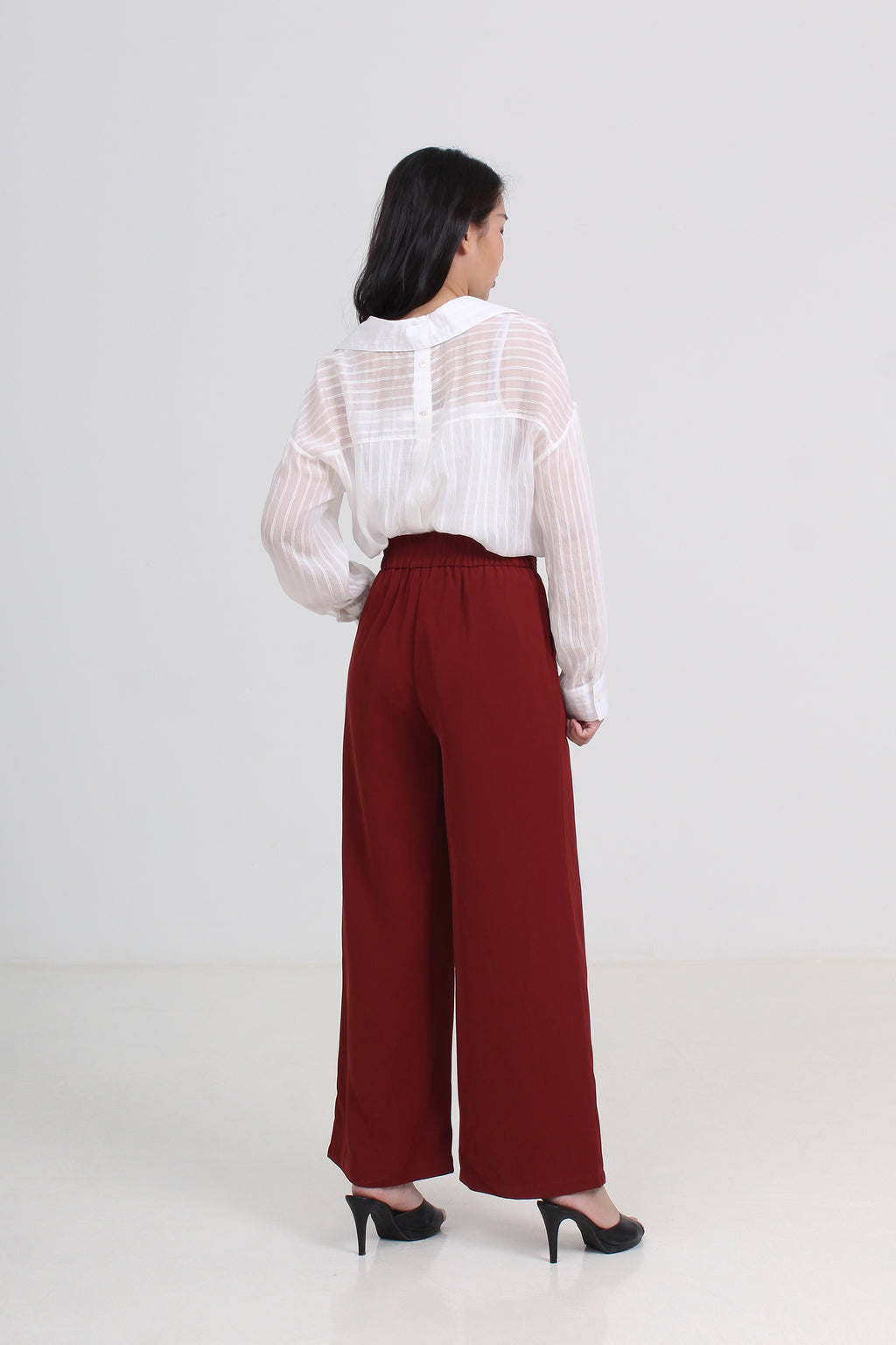 A woman is shown from the back wearing a white sheer long-sleeve blouse and high-waisted, wide-leg burgundy trousers with black heels