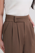 A close-up of a model wearing high-waisted brown pleated trousers with a matching pleated waistband, paired with a white ribbed top by le ann maxima.