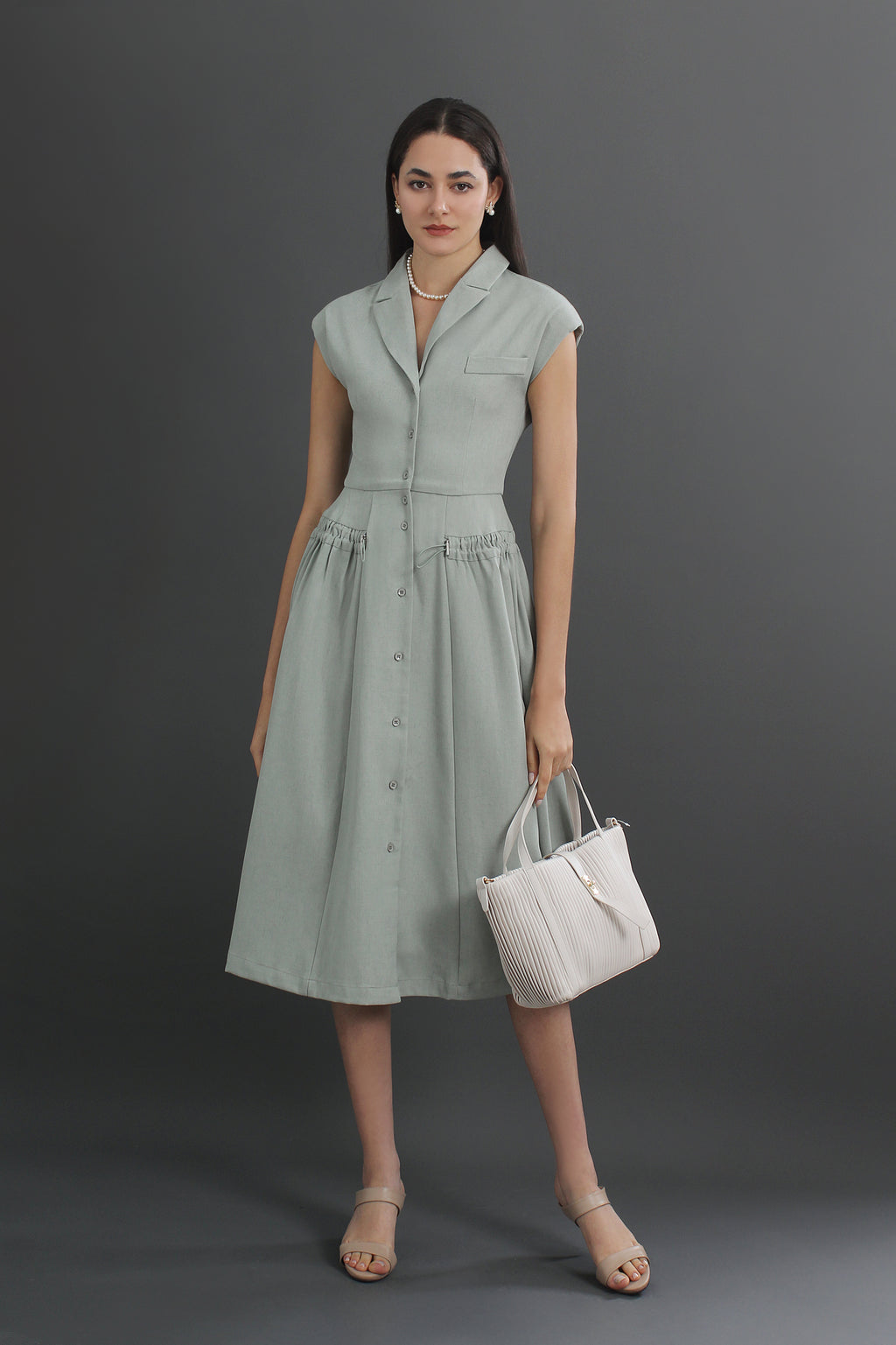 A woman standing and wearing a green V-neck linen collared midi dress with a pleated skirt, carrying a white tote bag.