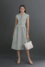 A woman standing and wearing a green V-neck linen collared midi dress with a pleated skirt, carrying a white tote bag.