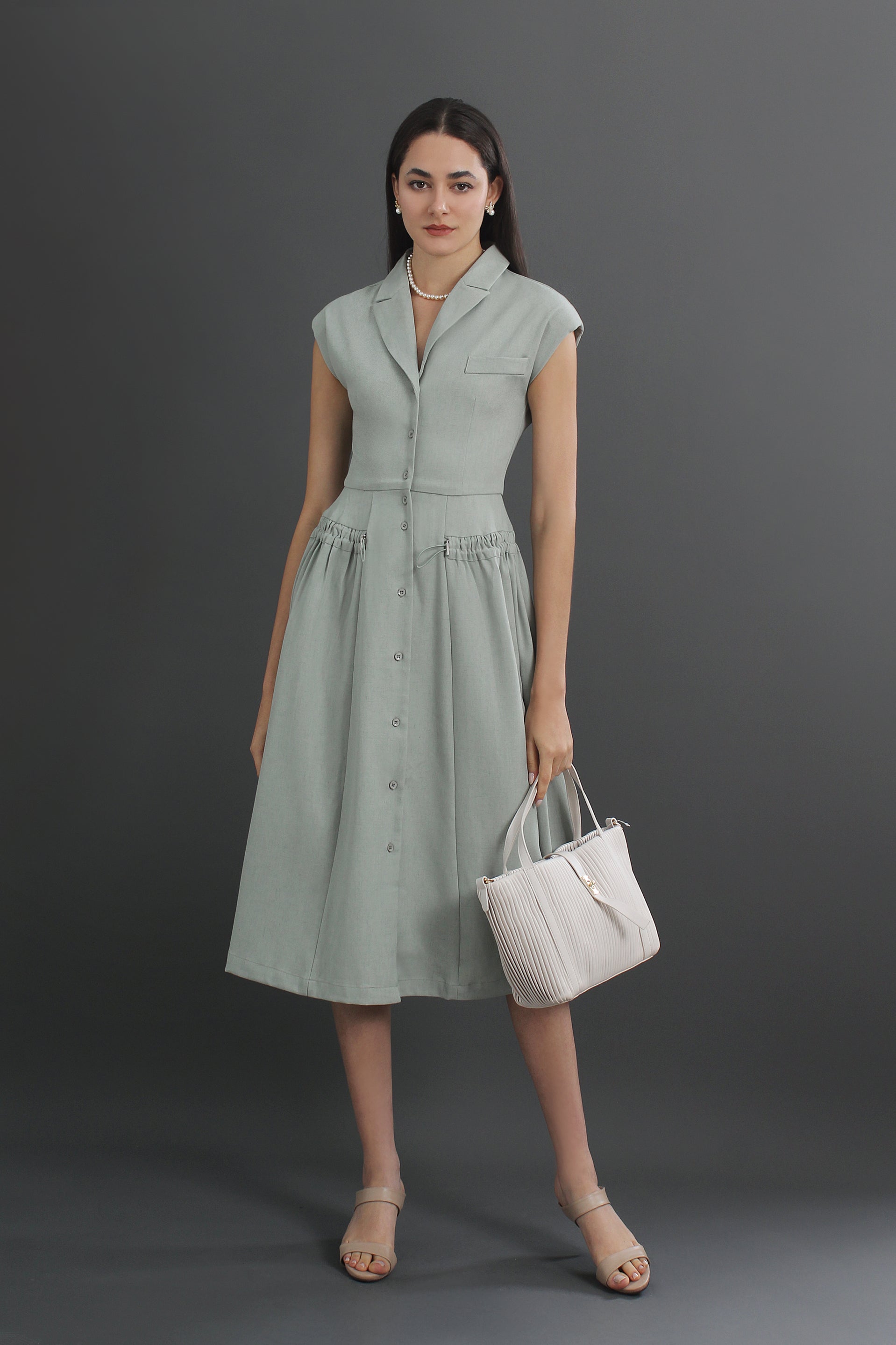 A woman standing and wearing a green V-neck linen collared midi dress with a pleated skirt, carrying a white tote bag.