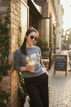 Woman wearing a grey snoopy and friends sweatshirt holding a drink outside a cafe with brick wall and greenery.