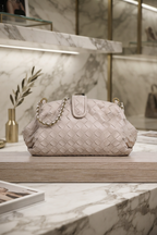 A woven leather handbag in a pale blush pink tone, featuring a gold chain strap and a central gold-tone hardware accent