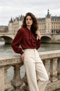A woman stands on a stone bridge in front of a large historic building over water, wearing a burgundy ruffled blouse and cream wide-leg trousers.