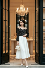 A woman wearing a black off-the-shoulder embellished top with long sheer sleeves, a white midi skirt